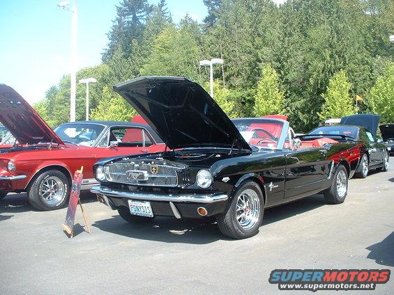 Black 1965 Ford Mustang convertible with hood open, red interior. Red Mustang and black car behind it