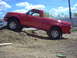 2001 Mazda B3000 - This red pickup truck is shown driving up a dirt mound under a partly cloudy sky.