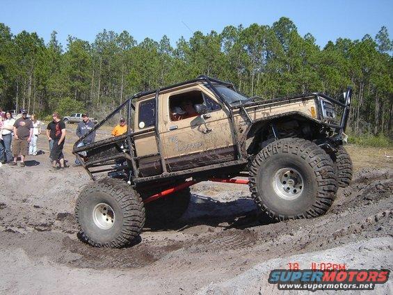 Mud-covered tan off-road truck with roll cage, large tires, and lifted suspension on a dirt track