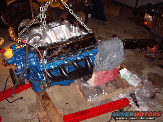 Blue V8 engine with chrome valve covers and black headers, attached to a gray transmission. Engine is suspended by chains