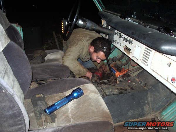 Man working on the floor of a vehicle interior, near the dashboard. Gloves and a flashlight are on the seat