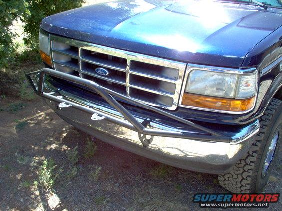 Blue Ford Bronco front end with chrome grille and aftermarket bumper. Tire and headlight visible