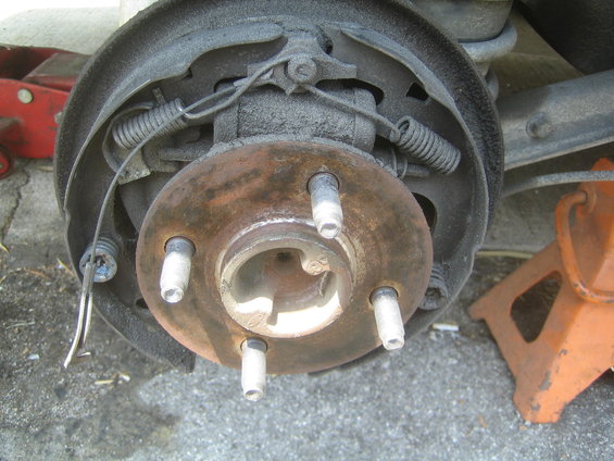 Rusty rear drum brake assembly with four lug nuts visible, supported by an orange jack stand
