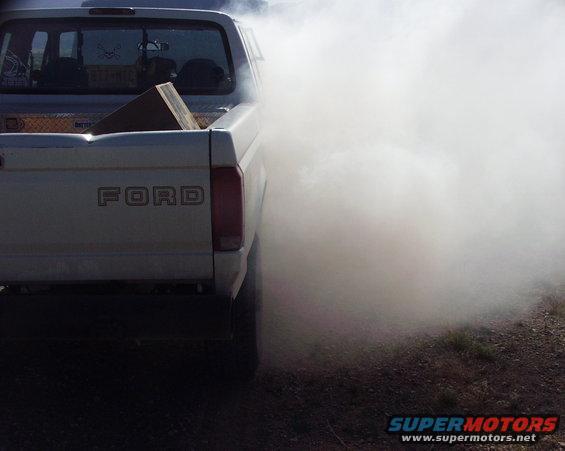 White 1997 Ford F-250 pickup truck emitting thick white smoke from exhaust, rear view
