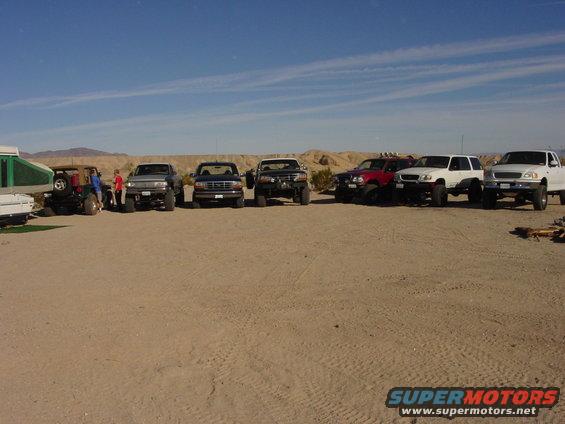 Several trucks and a Jeep parked in a desert lot with a pop-up camper