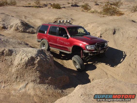 Red SUV with off-road modifications driving on sandy terrain. It has a roof rack with lights and large tires