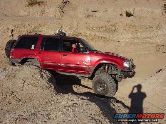 Red SUV with off-road modifications climbing a dirt hill. Photographer's shadow visible
