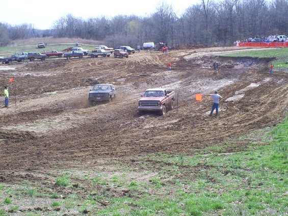 Red pickup truck and dark SUV racing on muddy track with spectators in background
