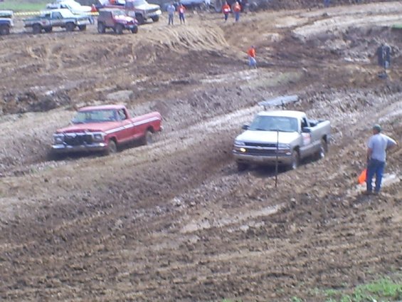 Two pickup trucks driving through mud during a race. One is red, the other is silver