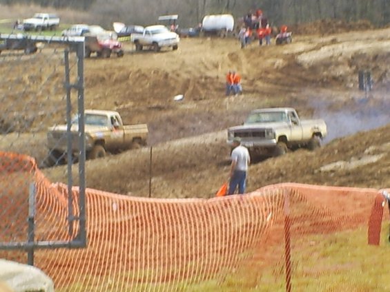 Two pickup trucks in a mud race, one in the foreground with a man standing nearby