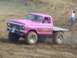 2001 Dodge 1500 - A pink flatbed pickup truck with oversized tires navigates a dirt track, with a person holding a flag in the background.