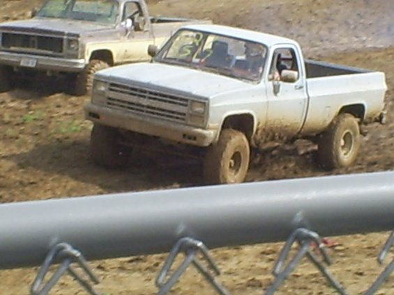 White pickup truck driving through mud, another older pickup behind it