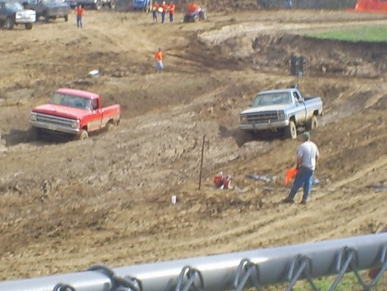 Silver pickup truck navigating a muddy track, with a red truck behind