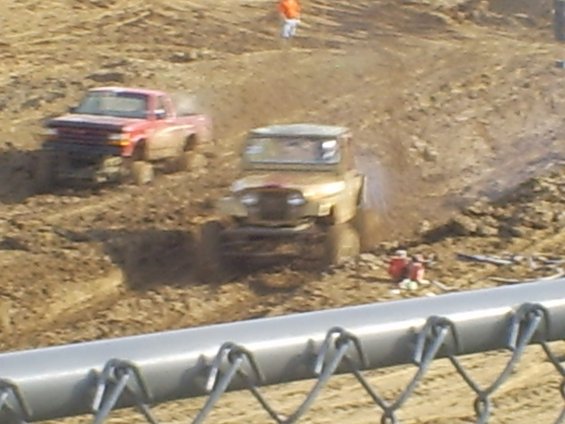 Tan off-road vehicle splashing mud, with red truck behind