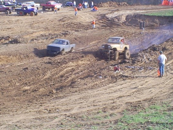 Mud bogging event with several off-road trucks and vehicles navigating a muddy track. One truck is stuck in the mud