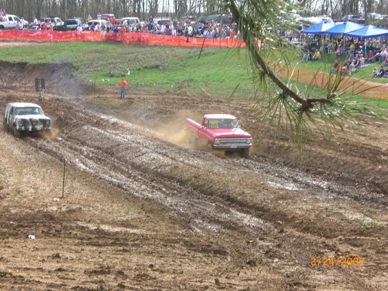 Red pickup truck kicking up mud during a race, with another white vehicle ahead. Spectators watch from the sidelines
