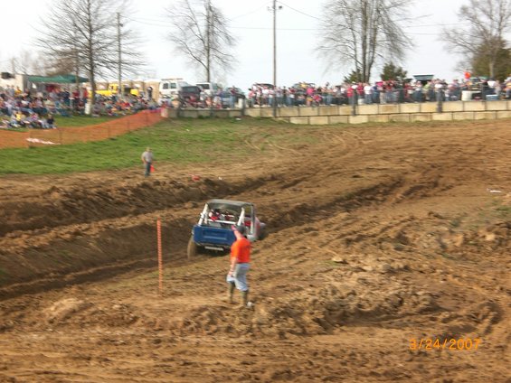 Blue off-road buggy going through a mud track with spectators in the background