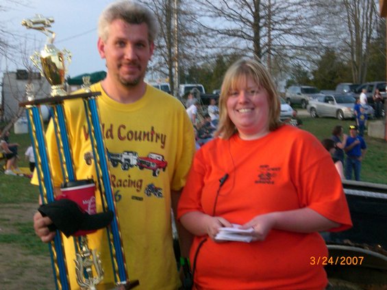 Man holding trophy, woman next to him, outdoor event with cars in background