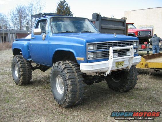Blue pickup truck with oversized tires and a white bull bar, parked on dry grass