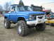 1990 Ford Bronco - A lifted blue pickup truck with large off-road tires sits on a grassy lot next to a trailer and another truck.
