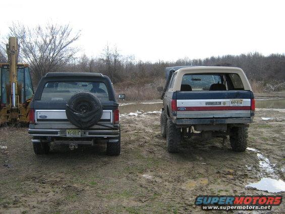 Two Ford Broncos, one black and one blue/silver/red, parked in a muddy outdoor area. A yellow backhoe is visible to the left
