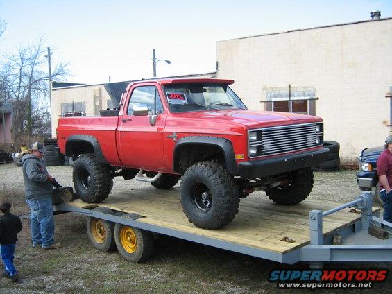 Red pickup truck with large tires on a trailer. "FOR SALE" sign in window