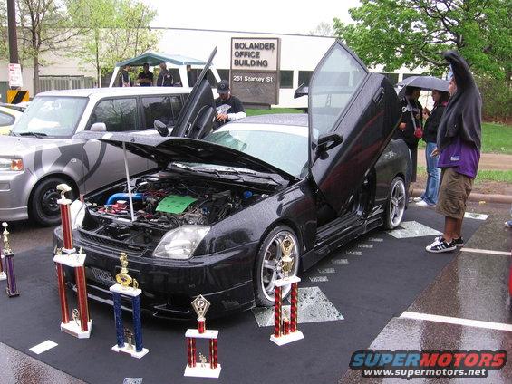 Black coupe with butterfly doors open, engine exposed, parked next to a silver SUV. Trophies are displayed in front of the coupe