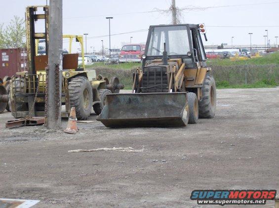 Yellow backhoe loader with front bucket and cab, next to a yellow forklift. Construction site setting
