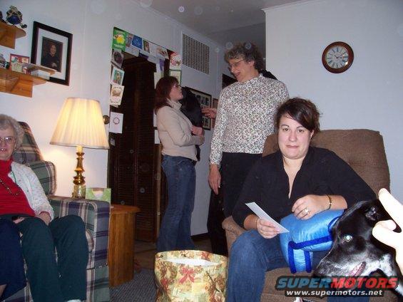 Four people and a dog are in a living room. A woman sits in a chair holding papers