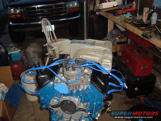 V8 engine block painted blue, with silver intake manifold and blue spark plug wires. Black Ford truck visible in background