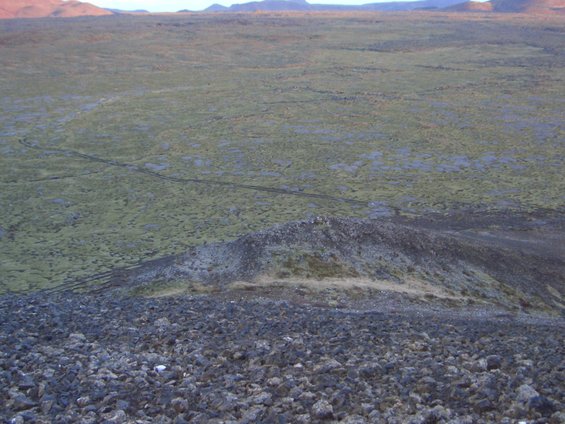 Rocky foreground slopes down to a vast, green, moss-covered plain with tire tracks, under a hazy sky