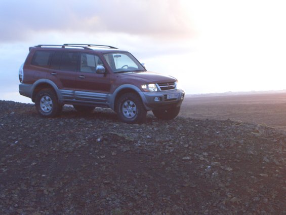 Red SUV with roof rack on rocky ground, license plate visible