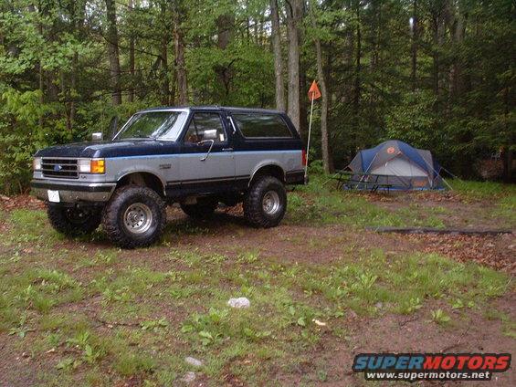 Two-tone blue and silver Ford Bronco with large tires parked on a dirt road next to a tent