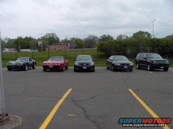Five cars parked in a lot: a black Corvette, a red Camaro, a dark sedan, another dark sedan, and a black SUV