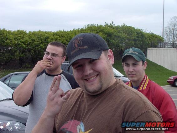 Three men stand outside, two in foreground, one in background. A car is visible on the left