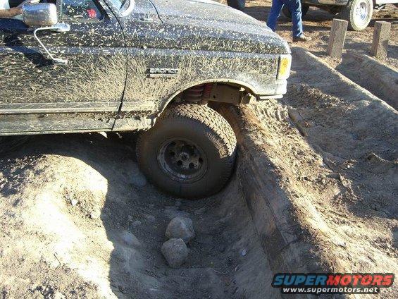 Muddy 1987 Ford Bronco going over an obstacle on an off-road course. Another vehicle is visible in the background