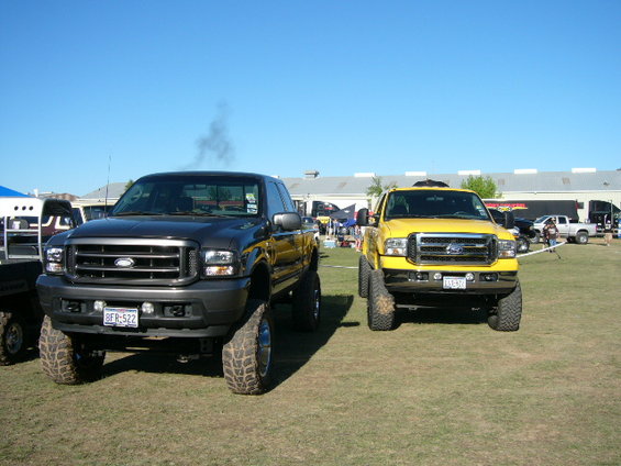 Two lifted Ford trucks, one gray and one yellow, parked on grass