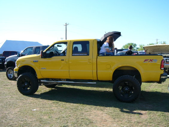 Yellow pickup truck with lifted suspension, people in the bed. Another truck is parked nearby