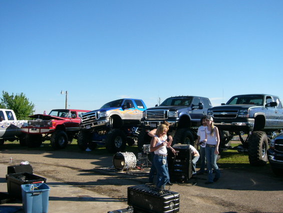 Several lifted pickup trucks are parked in a lot with people standing around them. Some trucks have their hoods up