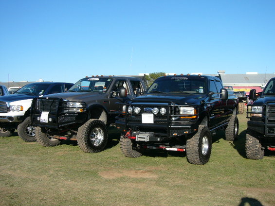 Several lifted Ford trucks parked on grass under a clear blue sky