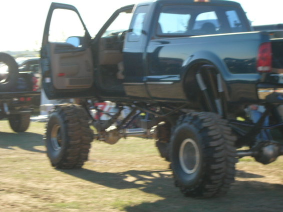 Lifted dark green pickup truck with open driver's door and large off-road tires. Another truck is visible to the left
