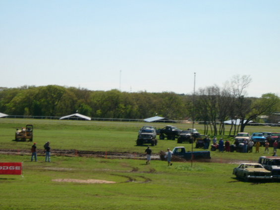 Several lifted trucks and SUVs gathered on a grassy field with a tractor and a classic car