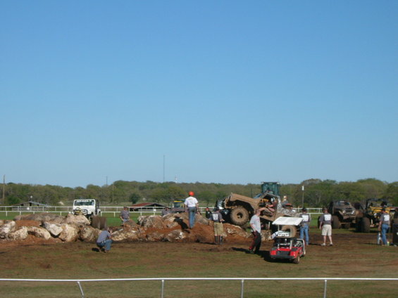 Off-road vehicles and people on a dirt and rock obstacle course under a clear blue sky