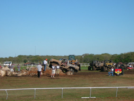Several off-road vehicles are in a muddy area with people around. A bulldozer is visible in the background