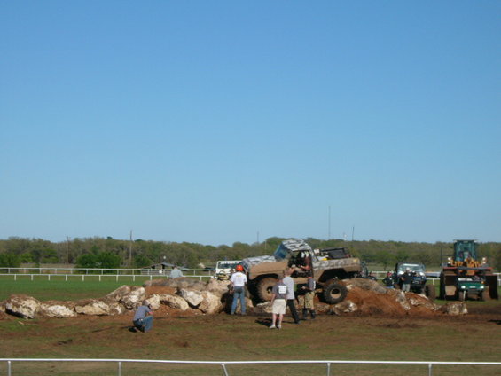 1978 Ford Bronco climbing over rocks with people watching. Another vehicle and a tractor are visible in the background
