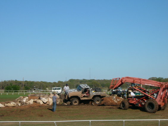 Camouflaged off-road truck navigating a rocky dirt course with a large red loader nearby