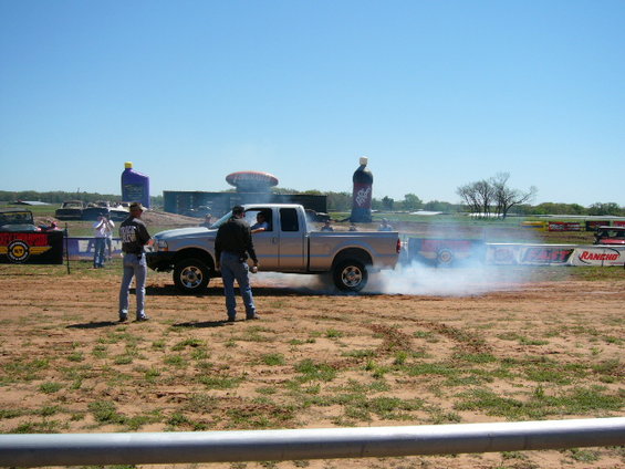 Silver pickup truck doing a burnout, kicking up smoke. Two men stand nearby watching