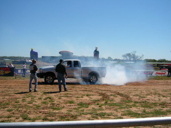 Pickup truck doing a burnout, kicking up a lot of smoke. Two guys are standing nearby watching