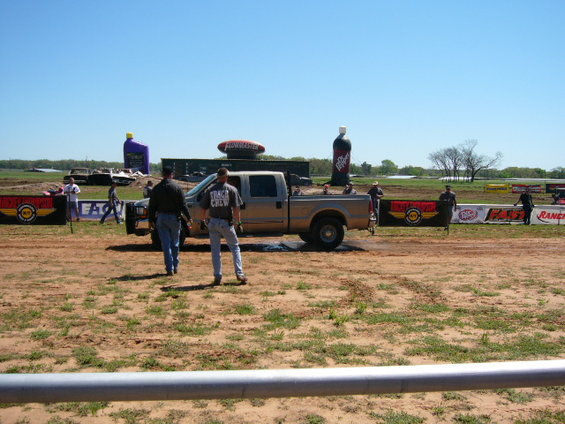 Pickup truck parked at a drag strip with two men walking away from it. Inflatable bottles and banners are visible
