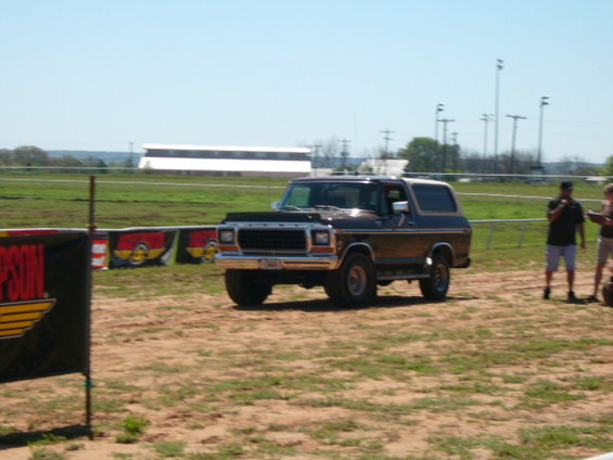 Brown 1978 Ford Bronco driving on dirt with two people standing nearby